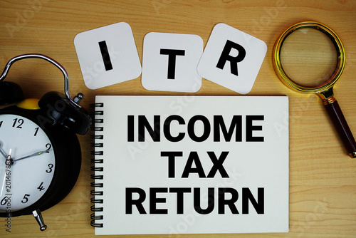 ITR Income Tax Return text written on notepad with magnifying glass and alarm clock on wooden background