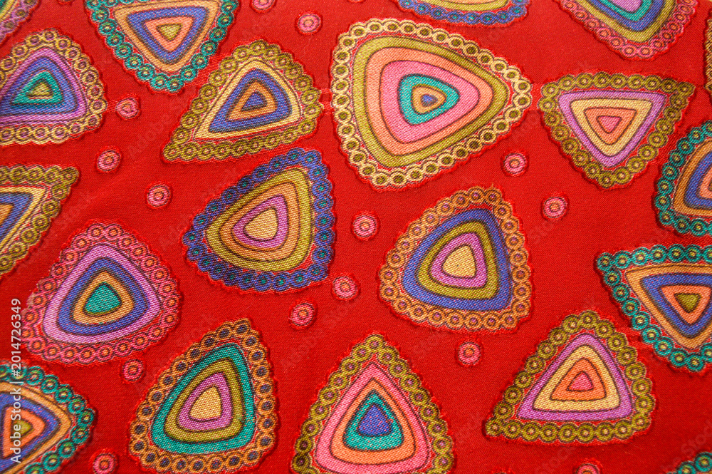 Fototapeta premium Close up of red fabric with colorful embroidered triangular patterns. Abstract textile background featuring vibrant geometric shapes and circular dot details.