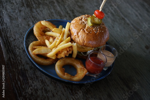 Delicious burger and fries served on a plate. The burger is topped with a pickle and tomato, accompanied by sauces, and onion rings.