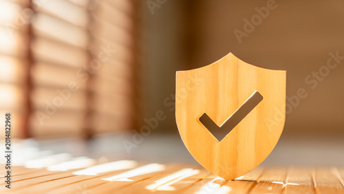Quality assurance and protection concept. Wooden shield with checkmark icon on desk under sunlight. Ideal for internet security, data privacy, health insurance, warranty and certified compliance.
