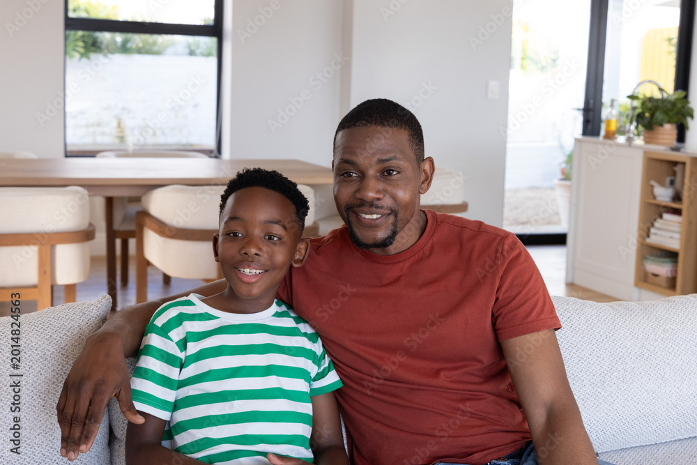 Fototapeta premium African American father and son sitting on upholstered couch in modern living-dining area smiling