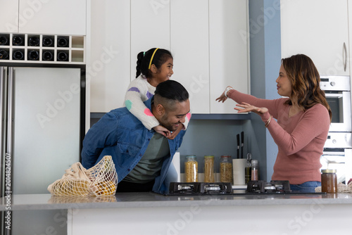 Family carrying child on father's back while mother gesturing at modern kitchen island with cooktop