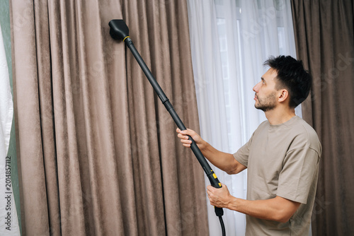 Fotografie Portrait of young bearded man professionally steam cleaning curtains in modern home, providing deep drapery maintenance and effective dry cleaning for textile hygiene and fresh living space