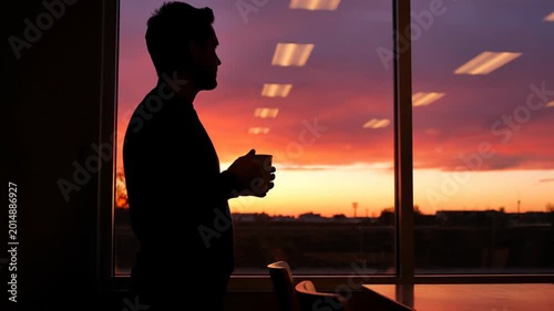 Silhouette of a man drinking coffee while looking out the office window at sunset.