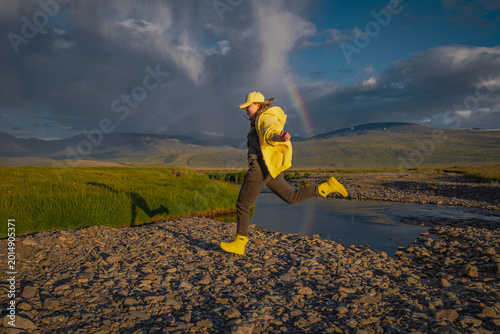 Foto Energetic young woman in yellow boots jumping over a stream under a rainbow and