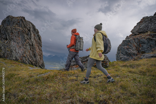 Fotografie Two determined hikers with backpacks and trekking poles walking past massive roc