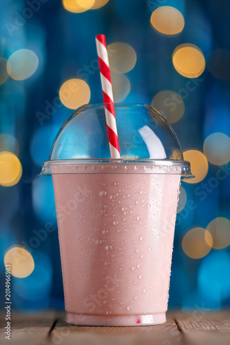 Chilled strawberry milkshake in disposable plastic cup with condensation and red and white striped straw on wooden surface, vibrant bokeh background, refreshing summer treat