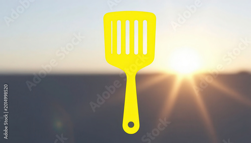A bright yellow spatula is prominently displayed against a sunset background. The spatula captures the light, emphasizing its color and shape during the sunset.