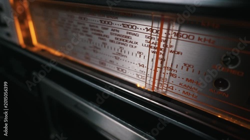 Close-up of an antique radio tuner dial glowing with warm orange light. The metallic panel displays various frequencies in kilohertz and megahertz. A red tuning needle rests on a station.