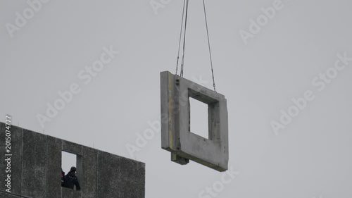 Construction scene unfolding. Steel cables hold precast panel near building facade. Worker observes as precast concrete panel is carefully hoisted into position