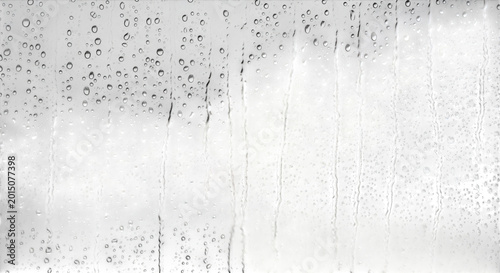 Raindrops and vertical streaks of water running down a transparent glass window pane against a blurred gray and white cloudy sky background