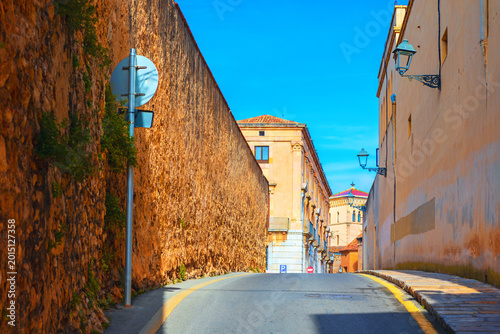 Tarragona showcases a narrow road flanked by an imposing ancient Roman stone wall and traditional residential buildings. Bright sunlight casts long shadows across the pavement leading ahead old town