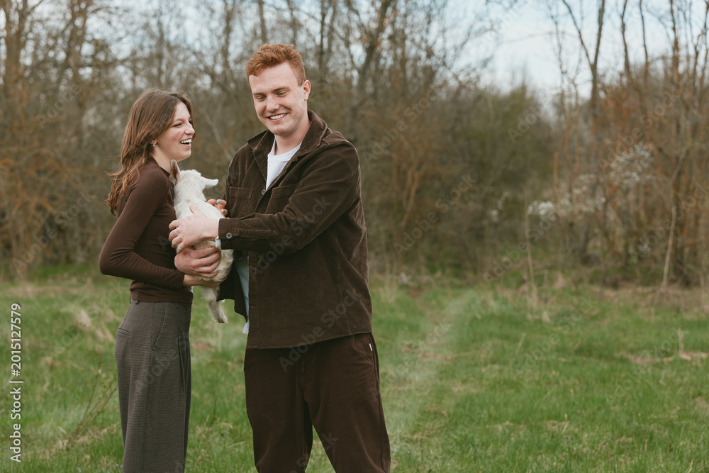 Fototapeta premium Joyful couple outdoors holds a newborn goat kid, smiling at each other, casual attire, in a grassy field with trees, warm natural light, candid lifestyle moment showcasing rural charm and connection