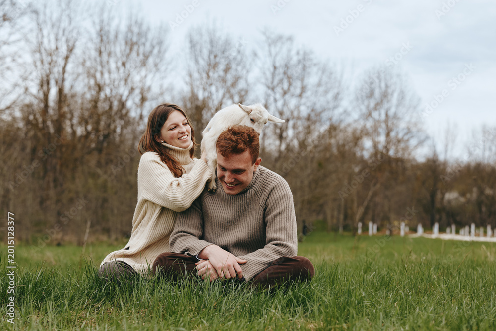 Fototapeta premium Cheerful couple outdoors, close in with a playful goat kid by their side, cozy sweaters, sunlit meadow, natural colors, relaxed posture, candid moment, nature backdrop, lifestyle portrait