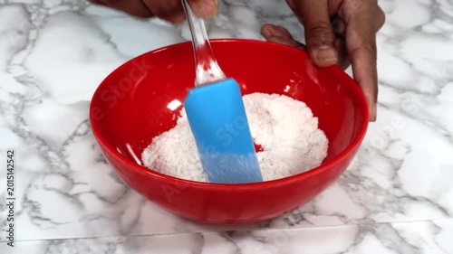 Stirring Dry Baking Ingredients in Red Bowl with Blue Spatula on Marble Countertop, Close-up of hands stirring dry baking ingredients in a bright red mixing bowl using a blue spatula