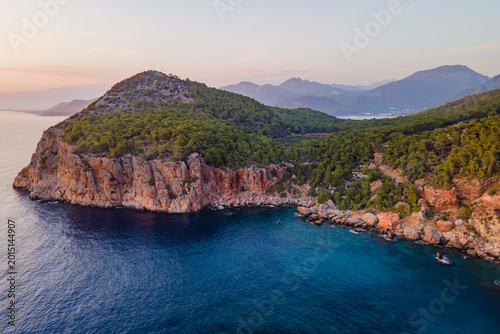 Aerial view of rocky Mediterranean coastline with forested hills and clear blue sea in Antalya region, Turkey. Coastal landscape at sunset with mountains and natural shoreline