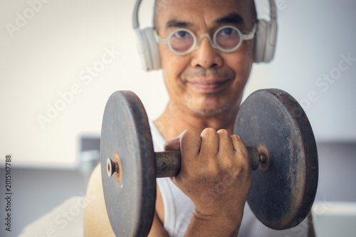 Happy senior Asian man exercising at home with dumbbells. Elderly male in white tank top and headphones enjoys lifting weights. Concept of active aging, healthy lifestyle, and fitness for retirees.