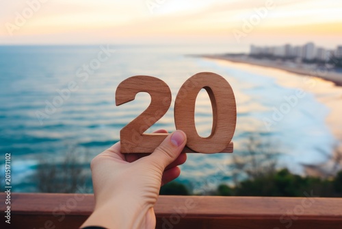 Hand holding wooden number 20, symbolizing celebration or anniversary against a coastal sunset