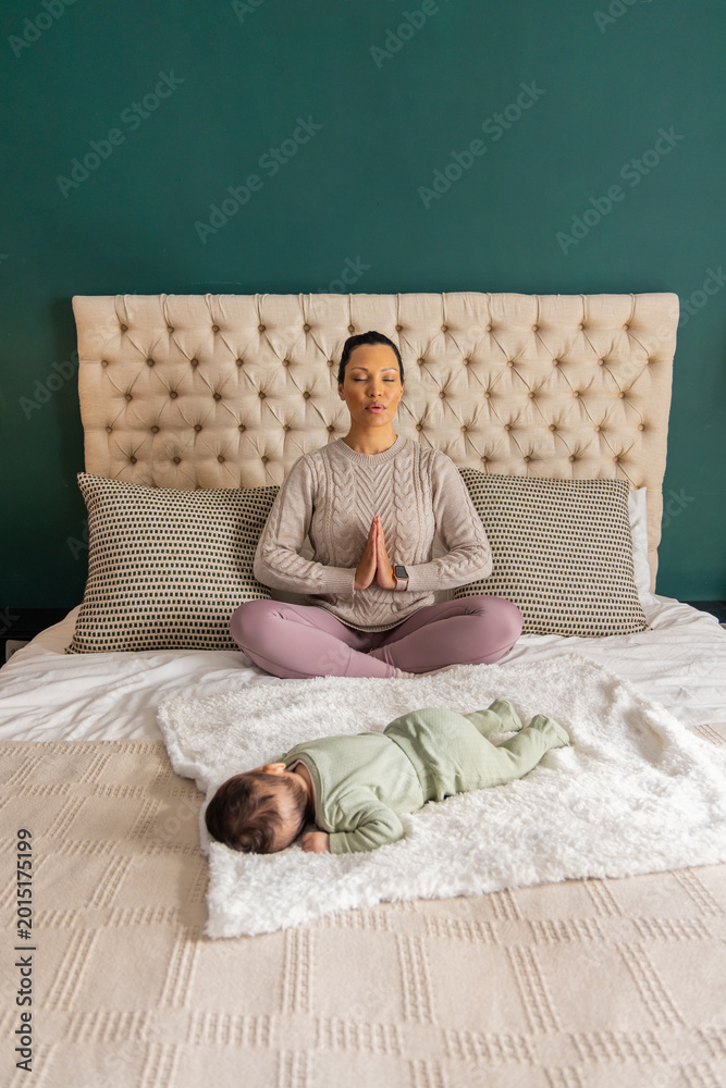 Fototapeta premium Mother sitting meditating with hands pressing on chest on bed wearing knit sweater, baby on blanket