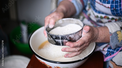 Obraz na plátně Elderly woman sifting flour through a mesh strainer, hands closeup showing flour