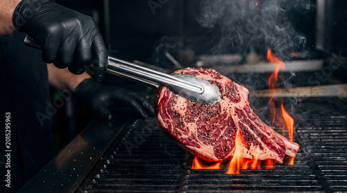 Chef grilling raw ribeye steak with tongs over open flame on dark grill