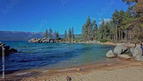 Sandy beach at Lake Tahoe with clear blue water and rocks in California. Scenic lakeshore landscape with pine forest and mountains, ideal for travel, nature, and outdoor use