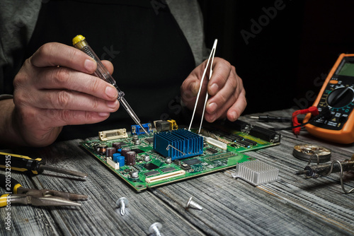A man uses a screwdriver and tweezers to repair a circuit board on a wooden table. Tools and a multimeter are also present in a low light setting