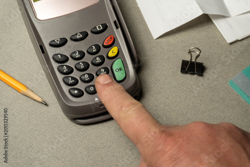 A finger presses the button on a payment device while office supplies like a pencil and binder clip sit on the grey table. Papers are around the device during the transaction