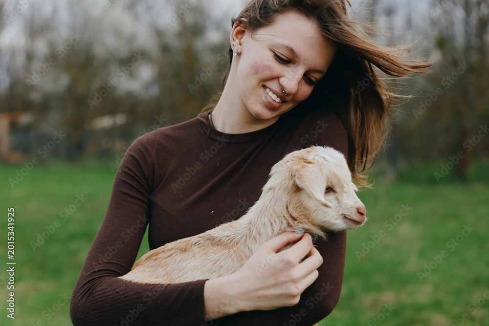 Fototapeta premium Happy woman in a brown sweater holds a playful goat kid in a sunlit field, sharing a tender moment outdoors, showcasing gentle animal affection amid open nature and rural countryside