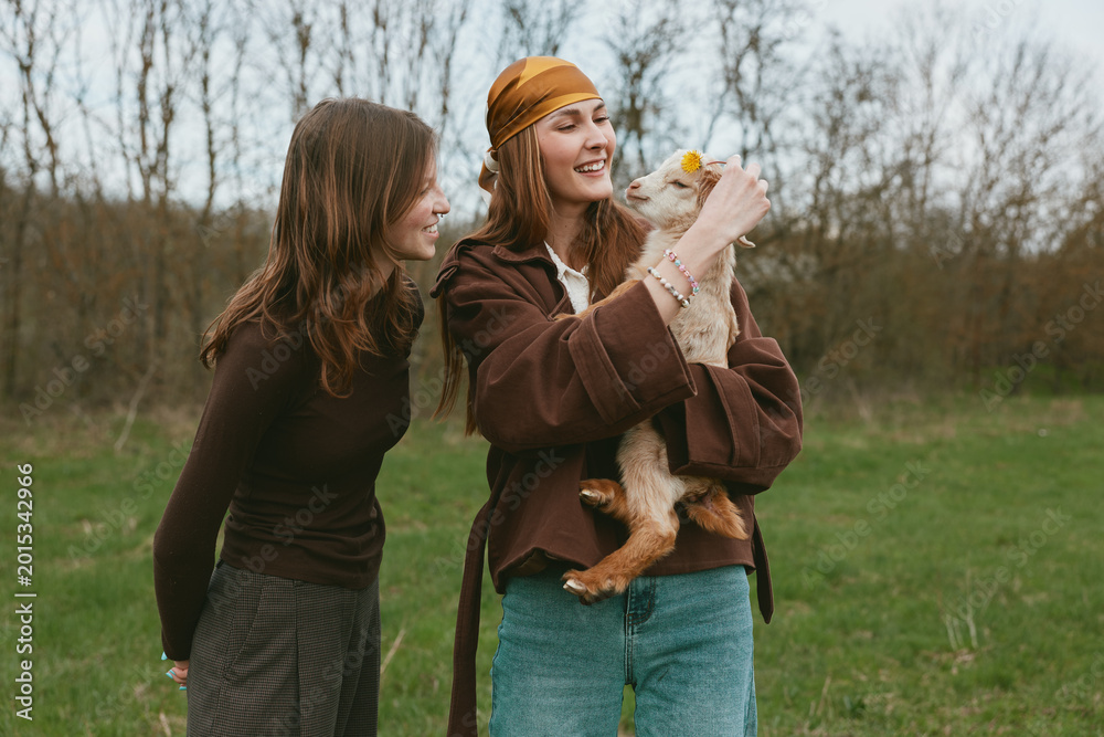 Naklejka premium Two women in casual clothes share a joyful moment with a goat kid in a grassy meadow, outdoor. A warm lifestyle scene highlighting bonding, care, and rural life.