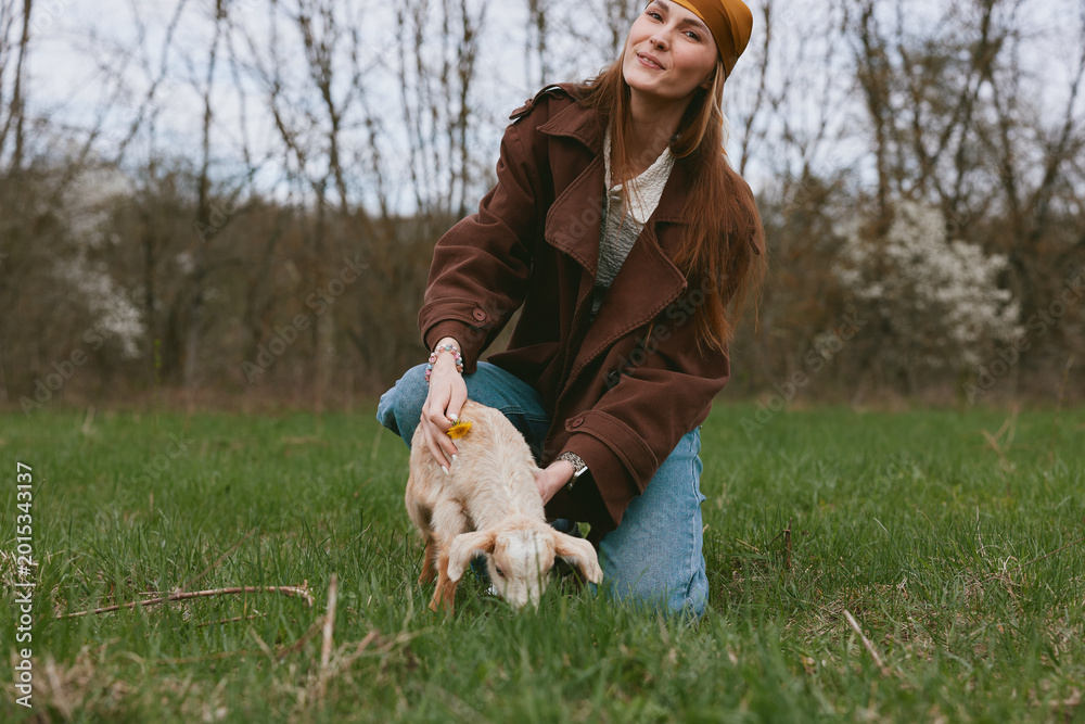 Fototapeta premium Friendly woman in a brown coat kneels on green grass, guiding a playful goat kid. Outdoor lifestyle scene with natural light, showing care, companionship, and a rustic countryside mood.