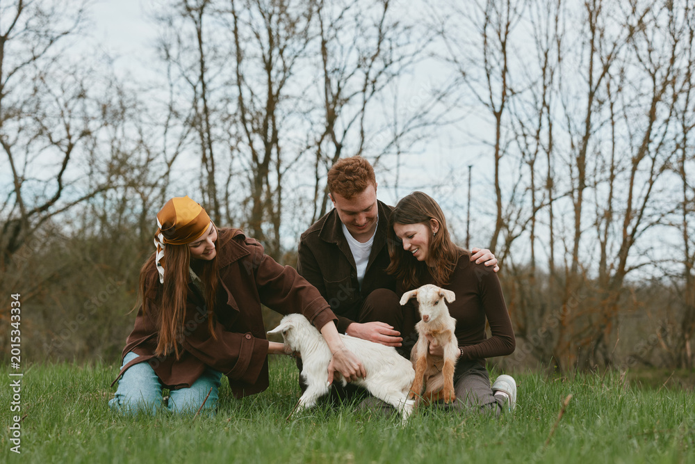 Fototapeta premium Three young adults enjoy a casual outdoor moment with two baby goats in a grassy field during early spring, capturing a warm and natural lifestyle scene.