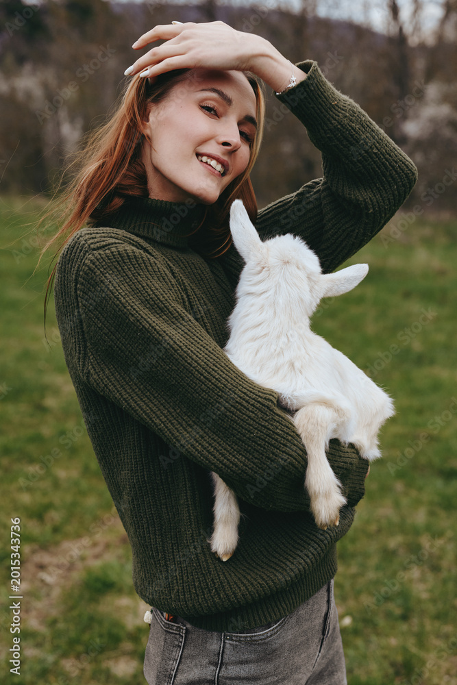 Fototapeta premium woman goat kid outdoors lifestyle joyful smiling sweater field countryside sunlit nature affection cuddle animal companion rural grass meadow, a warm, candid moment captured with natural colors and