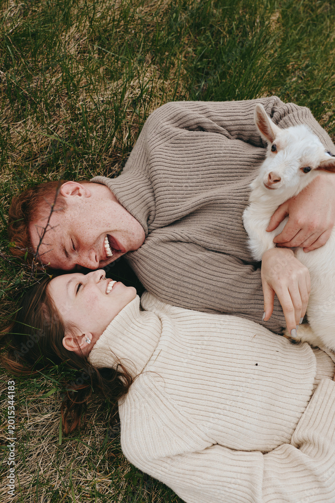 Obraz premium Warm lifestyle scene with a smiling couple on the grass holding a goat kid, cozy sweaters, and natural light creating a tender, carefree mood in a peaceful outdoor setting