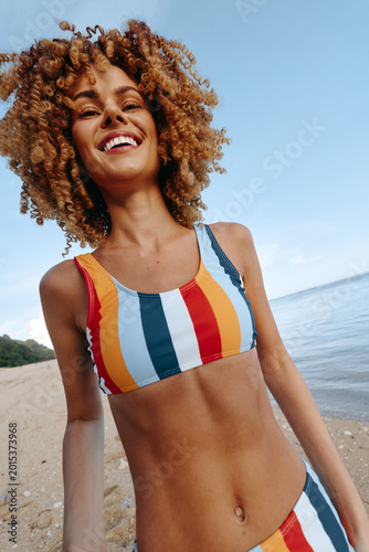 A smiling woman at the beach wears a colorful striped bikini, capturing a carefree portrait by the sea with blue sky and bright sunlight, embodying summer vibes.