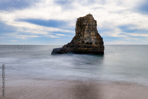 Isolated sea stack rising from calm Pacific Ocean, long exposure waves creating soft motion around rocky formation under cloudy sky along California coast