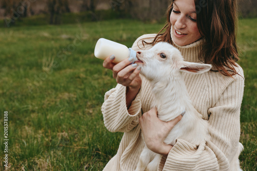 A woman in a soft sweater gently bottles a hungry kid in an open field, capturing a tender moment of farm life, nurture, and animal care in a bright outdoor setting.