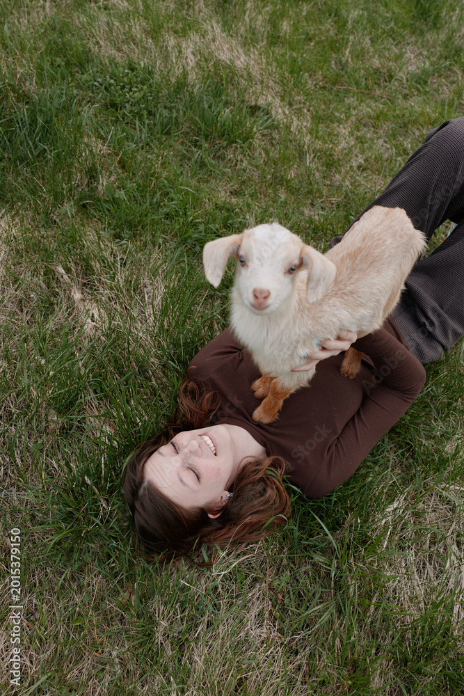 Obraz premium A young woman lies on grass smiling as she holds a small white baby goat on her chest in a relaxed outdoor lifestyle scene.
