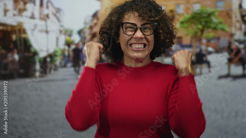 Foto Woman wearing red sweater clenches fists amid outdoor cafe seating on city street under warm sunlight; excitement
