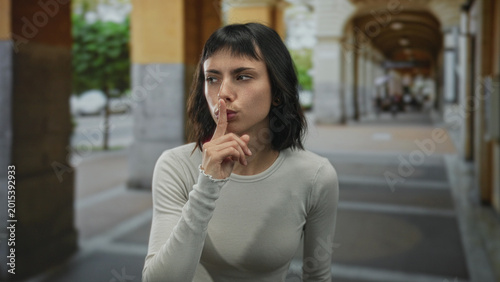 Фотография Young hispanic woman finger to lips for silence while standing on sunlit street colonnade beneath historic building arches; secrecy