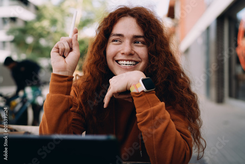 Excited red-haired model holding stylus and looking away with big smile. Successful creative idea, brainstorming session, remote student rejoices, happy freelancer in outdoor cafe, inspiration.