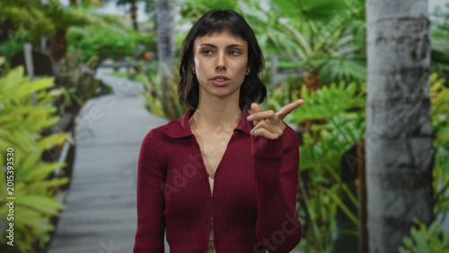 Photos Woman in red cardigan points finger to herself on a street walkway surrounded by green foliage; refusal defiance