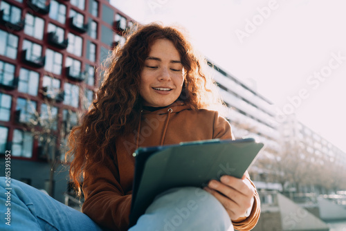 Happy red-haired student looking at digital tablet outdoors against city architecture. Remote education process, preparing for exams, online research, creative writing, digital nomad lifestyle.