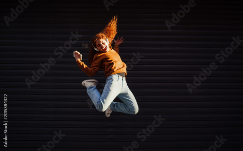 Quadro su tela Cheerful redhead man with smartphone jumping high against dark wall, celebrating victory, success and achievement, active digital nomad, joy emotion, authentic Caucasian model person