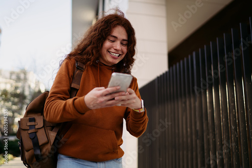 Cheerful ginger man with backpack using mobile phone outdoors, laughing at screen content, social media messaging, digital nomadic life, 5G internet connection, Caucasian model, real people.