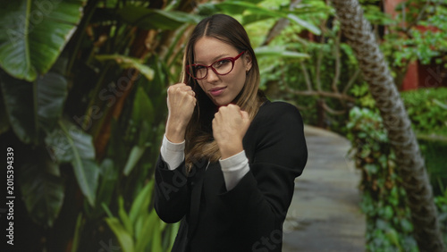 Fotografia Woman standing with fists raised and red glasses on a sunlit forest path among lush green plants; determination confidence resilience