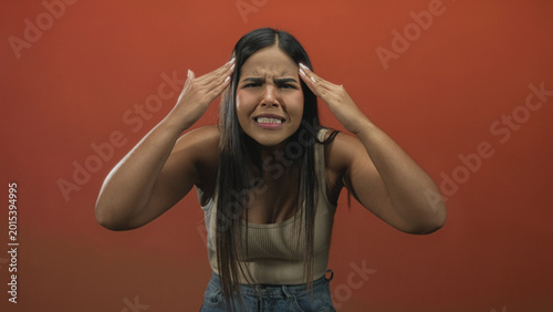 Billede på lærred Hispanic woman with raised arms and pointed finger gesturing accusation in studio wearing tank top and jeans against red backdrop; frustration