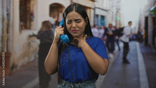 Billede på lærred Young woman holding a blue telephone handset to her ear and clutching her neck on a busy urban street lined with storefronts and blurred pedestrians; panic plea