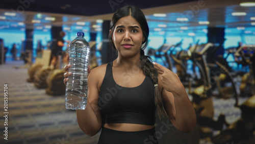 Foto Young hispanic woman in black sports bra holds a large water bottle and points to herself in a gym; hydration confusion