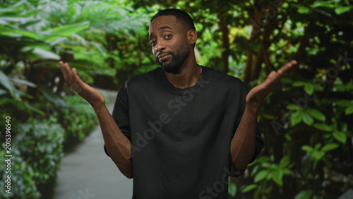 Photos Man in black t shirt shrugs with palms up on a leafy green park path, facing camera with a slight smile; uncertainty casual relaxed
